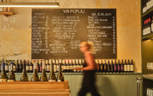 The interior of Vin Populi features a blackboard with their Italian food menu items and their wine list. Underneath, a sidetable is lined with bottles of wine