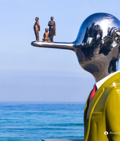Metal sculture of a man with a long nose and suit and a small family on his nose in front of the ocean Image